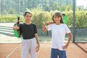 barn och sporter begrepp. porträtt av leende flickor Framställ utomhus- på padel domstol med racketar och tennis bollar foto