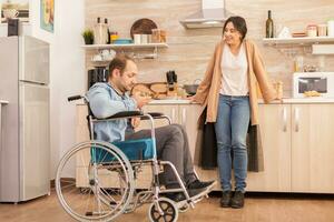Inaktiverad man i rullstol ser på sallad tillverkad förbi fru i kök. Inaktiverad lam handikappade man med gående handikapp integrering efter ett olycka. foto