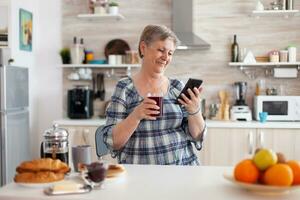 senior kvinna använder sig av mobil grej i de kök dricka aromatisk te i de morgon- under frukost. äkta äldre person sökande på modern smartphone internet teknologi, senior fritid tid foto