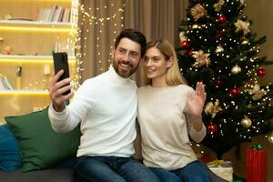 gift par man och kvinna tillsammans på soffa på Hem jul träd med smartphone talande video ring upp och tar selfie, mogna familj hälsning vänner uppkopplad avlägset för ny år och jul. foto