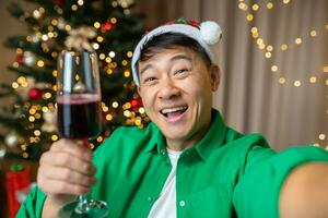 Lycklig ung stilig asiatisk man i röd santa claus hatt och grön skjorta tar selfie på Hem nära jul träd, innehav glas av vin, leende. foto
