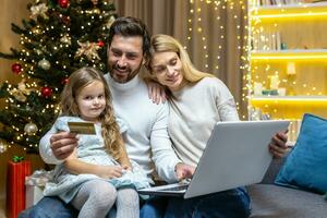 Lycklig jul familj håller på med uppkopplad handla Sammanträde på Hem på soffa, par Make fru och dotter med bärbar dator och Bank kreditera kort har roligt välja jul gåvor nära jul träd. foto