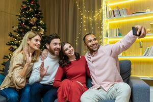 ny år fest Hem olika grupp av vänner, män och kvinnor, Sammanträde på de soffa, tar en selfie tillsammans, använder sig av en smartphone, gäster ser på de telefon, talande på en video ring upp för jul foto