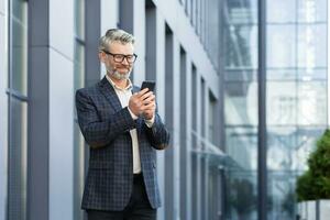 mogna vuxen affärsman utanför kontor byggnad gående och använder sig av smartphone, chef skriver meddelande i uppkopplad Ansökan på telefon, leende och Lycklig läsning Nyheter uppkopplad. foto