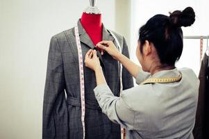 asiatisk kvinnlig modedesigner tjej som passar på den formella kostymen enhetliga kläder på skyltdocka modell. modedesigner eleganta showroom. sömnad och skräddarsy koncept. kreativ frisörstylist foto