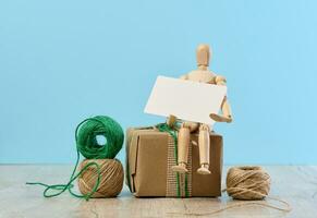 trä- mannekäng innehav en vit papper företag kort och gåvor insvept i brun hantverk papper på en blå bakgrund foto