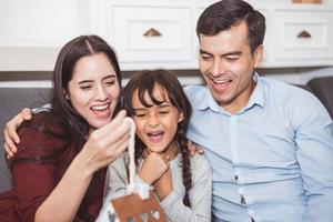 far och mor överraskar sin dotter med gåva eller ny leksak. föräldrar och barn är lyckliga tillsammans hemma på soffan i vardagsrummet. familj och livets lycka koncept