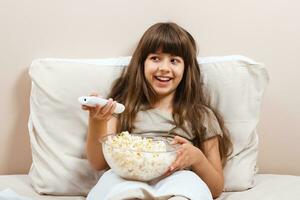 liten flicka är tittar på TV och äter popcorn i henne säng. foto