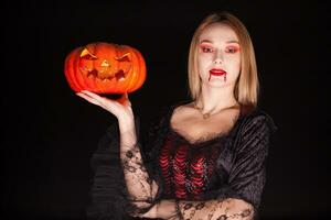 skön blond kvinna klädd upp tycka om ett ondska vampyr innehav en pumpa för halloween. foto