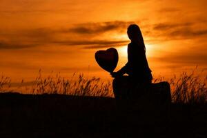 silhuett av en kvinna Sammanträde på resväska med hjärta formad ballong och ser på skön solnedgång. foto