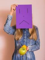 kvinna innehav frukt och ledsen ansikte på papper medan stående i främre av vägg. foto