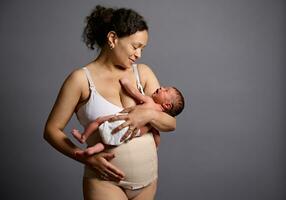 Lycklig mor i underkläder och elastisk bandage på henne mage efter c-sektion, leende ser på henne nyfödd bebis foto