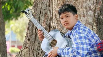 ung asiatisk pojke är spelar gitarr i en lokal- parkera, mjuk och selektiv fokus foto