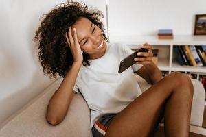 svart ung kvinna i hörlurar med mobiltelefon medan hon vilar på soffan