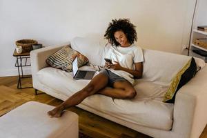 svart ung kvinna som använder mobiltelefon medan hon vilar på soffan foto