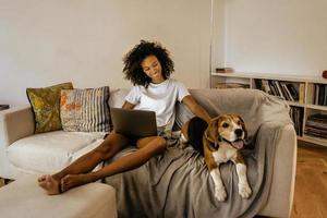 svart ung kvinna som använder bärbar dator och stryker hennes hund på soffan foto