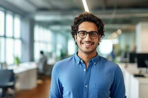 ai genererad en ung indisk manlig designer, ingenjör, arkitekt vem är bär glasögon och en blå skjorta leende stående i de kontor och ser på de kamera foto
