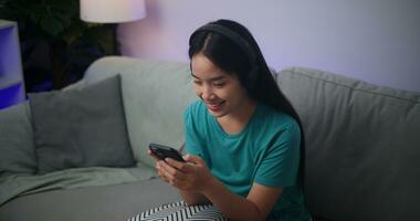 porträtt av ung asiatisk kvinna bär glasögon och hörlurar åtnjuter spelar uppkopplad esport spel på smartphone Sammanträde på soffa i de levande rum på hem, spelare livsstil begrepp. foto