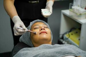 närbild kosmetolog använda sig av kosmetisk borsta applicering ansiktsbehandling gelé gel på kvinna klient ansikte i skönhet klinik. foto