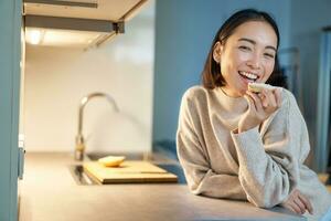 porträtt av leende ung Lycklig kvinna vistas på Hem, stående i kök och äter rostat bröd, ser åt sidan foto