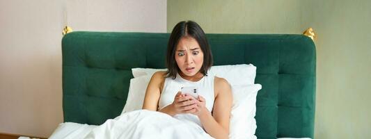 sovrum bild av asiatisk kvinna liggande i säng, ser rädd och chockade på smartphone skärm, läsning meddelande med bekymrad ansikte foto