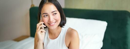 leende koreanska flicka i säng, samtal på mobil telefon, framställning en telefon ringa upp, lat morgon- som asiatisk kvinna order leverans via smartphone foto