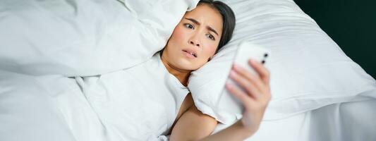 porträtt av asiatisk kvinna vakna upp i säng, ser chockade på mobil telefon, inse hon försovat foto