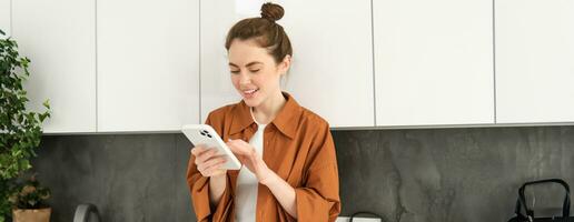 bild av skön ung kvinna modell, Framställ med smartphone i de kök, ser på mobil skärm och leende medan läsning en meddelande på telefon foto
