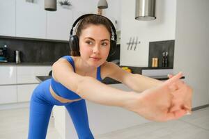 stänga upp porträtt av passa och friska kvinna, leende medan håller på med träna övningar, lyssnande musik i trådlös hörlurar, stretching vapen, aerobics Träning på Hem, bär blå aktiva kläder foto