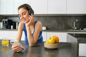 bild av Lycklig, eleganta ung sporter kvinna, stående i kök och dricka orange juice, lyssnande musik i hörlurar, använder sig av smartphone app foto
