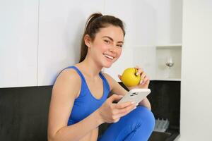 porträtt av brunett kondition kvinna, äter ett äpple, innehav smartphone, använder sig av mobil telefon app medan har friska frukt mellanmål i kök foto