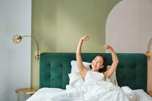 porträtt av leende Lycklig asiatisk flicka, vaknar upp känsla entusiastisk, sträckor henne händer upp, åtnjuter Bra morgon- i henne sovrum foto