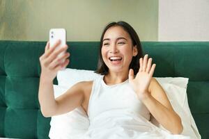 porträtt av leende koreanska kvinna ser på smartphone, liggande i säng och video chattar, vinka hand på mobil telefon kamera, tar selfie, inspelning video foto