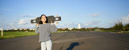 livsstil och människor. ung asiatisk flicka Framställ med longboard, skridskoåkning på henne kryssare. leende kvinna innehav skateboard på axlar foto