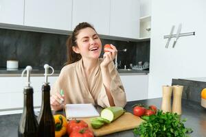porträtt av Lycklig brunett kvinna, biter tomat, sitter i de kök med grönsaker, matlagning vegetarian måltid, framställning lista av specerier, middag meny, bär morgonrock foto