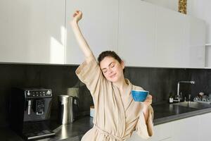 porträtt av Lycklig flicka dans med kaffe i de kök, bär morgonrock, njuter henne morgon- rutin- foto