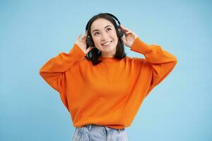 Lycklig kinesisk kvinna i hörlurar, lyssnar musik, åtnjuter favorit låt i henne Spellista, står över blå bakgrund foto