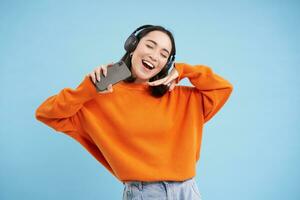 skön Lycklig koreanska kvinna i hörlurar, lyssnande musik på mobil telefon i hörlurar, bär orange tröja, blå bakgrund foto