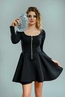 blond kvinna i svart kort klänning är innehav några kontanter, Framställ mot grå bakgrund. hasardspel underhållning, poker, kasino. närbild. foto