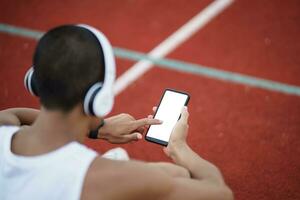 asiatisk man sportsman passa kropp bär vit hörlurar och använder sig av mobil telefon på vit skärm till lyssna musik medan vilar eller innan joggning, utövar på körfält stadion. sport friska löpning begrepp. foto