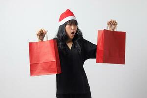porträtt av överraskad asiatisk kvinna med röd santa hatt känsla Lycklig medan köp av grejer. upphetsad flicka innehav handla väska. ny år och jul begrepp. isolerat bild på vit bakgrund foto