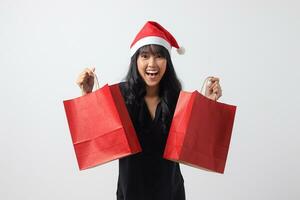 porträtt av attraktiv asiatisk kvinna med röd santa hatt känsla Lycklig medan köp av grejer. upphetsad flicka innehav handla väska. ny år och jul begrepp. isolerat bild på vit bakgrund foto
