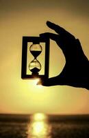 en person innehav upp en timglas i främre av de solnedgång foto