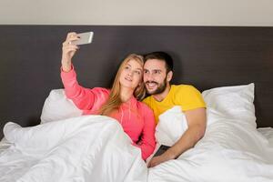 ung Lycklig par på säng håller på med selfie med telefon kamera. foto