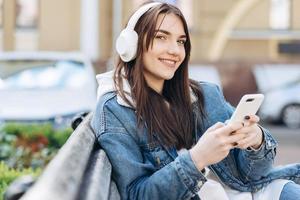 leende tjej som sitter på en bänk och njuter, lyssnar på musik i vita, trådlösa hörlurar, håller en smartphone i handen. gata med bilar på en suddig bakgrund foto