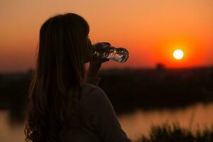 silhuett av en kvinna dricka vatten på de solnedgång. foto
