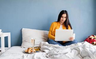 ung brunett kvinna som sitter i sängen och arbetar på dator som dricker champagne foto