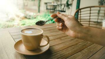 hjärta formad kaffe latte i en brun kopp med en fat, placerad på ett gammal trä- tabell. hand innehav en sked för bryggning kaffe. den där är redo till ge du friskhet varje morgon. foto