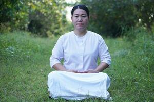 asiatisk kvinna buddist bär vit kostym, sitter och förbereda till do meditation på lugn skog. begrepp, kultur av tro. religiös aktivitet. fred av sinne förvaltning. göra koncentration. foto