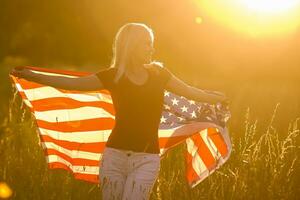 skön ung flicka innehav ett amerikan flagga i de vind i en fält av råg. sommar landskap mot de blå himmel. horisontell orientering. foto